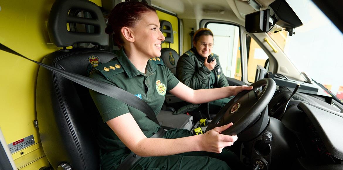 Two women in green uniform sitting in the front of an ambulance cab, with one steering with hands on the steering wheel and the other talking into the radio.