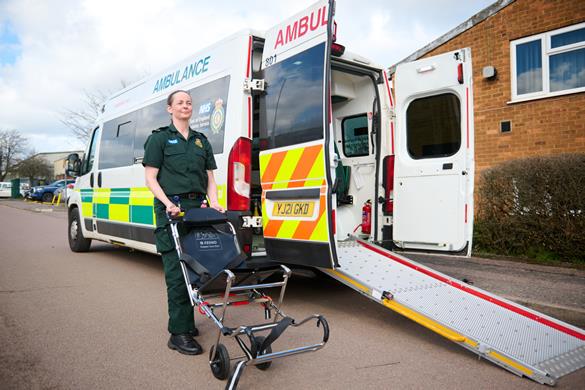 Non-emergency ambulance (PTS vehicle) with female crew member standing beside it pushing an empty carry chair.